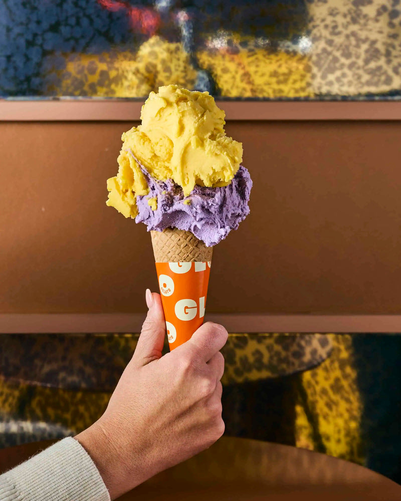 Hand holding a double-scoop gelato cone with yellow and purple flavors in a branded GLO Pasticceria sleeve