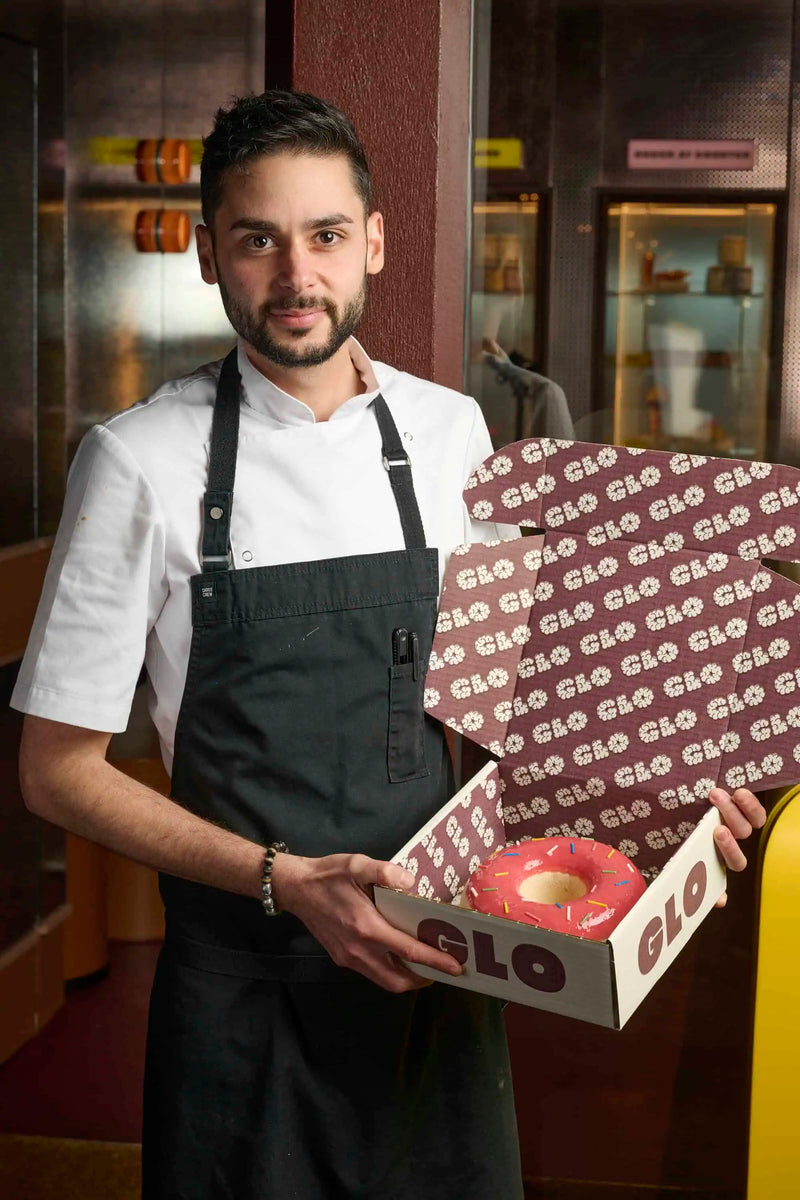 Chef holding a pink frosted donut in a branded GLO Pasticceria pastry box inside the bakery